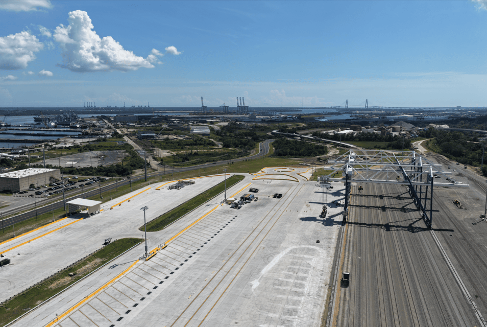 An expansive aerial view of a modern intermodal shipping terminal featuring massive gantry cranes and rail infrastructure under a clear blue sky.