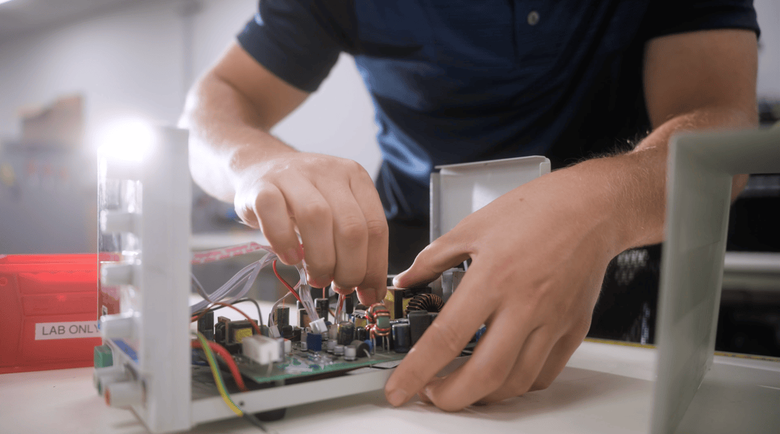 Close-up of a technician's hands working on the internal circuitry of an electronic device in a lab setting.