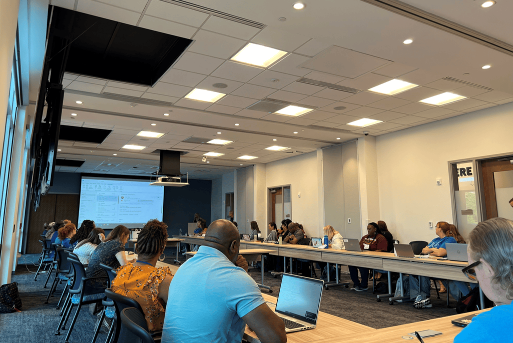 A group of adults seated around long conference tables in a modern meeting room, working on laptops and listening to a presentation projected on a screen at the front of the room