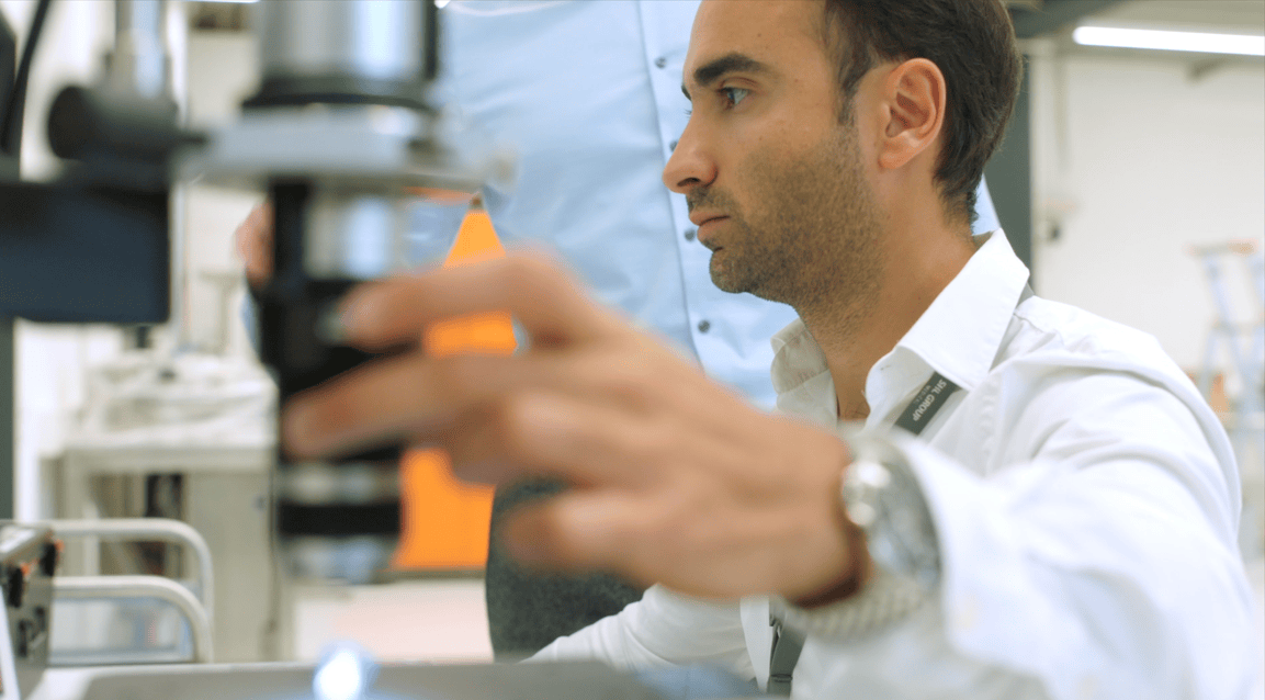 A male researcher wearing an SHL Group lanyard focuses intently while using his hand to adjust the lens of a professional microscope in a brightly lit lab setting