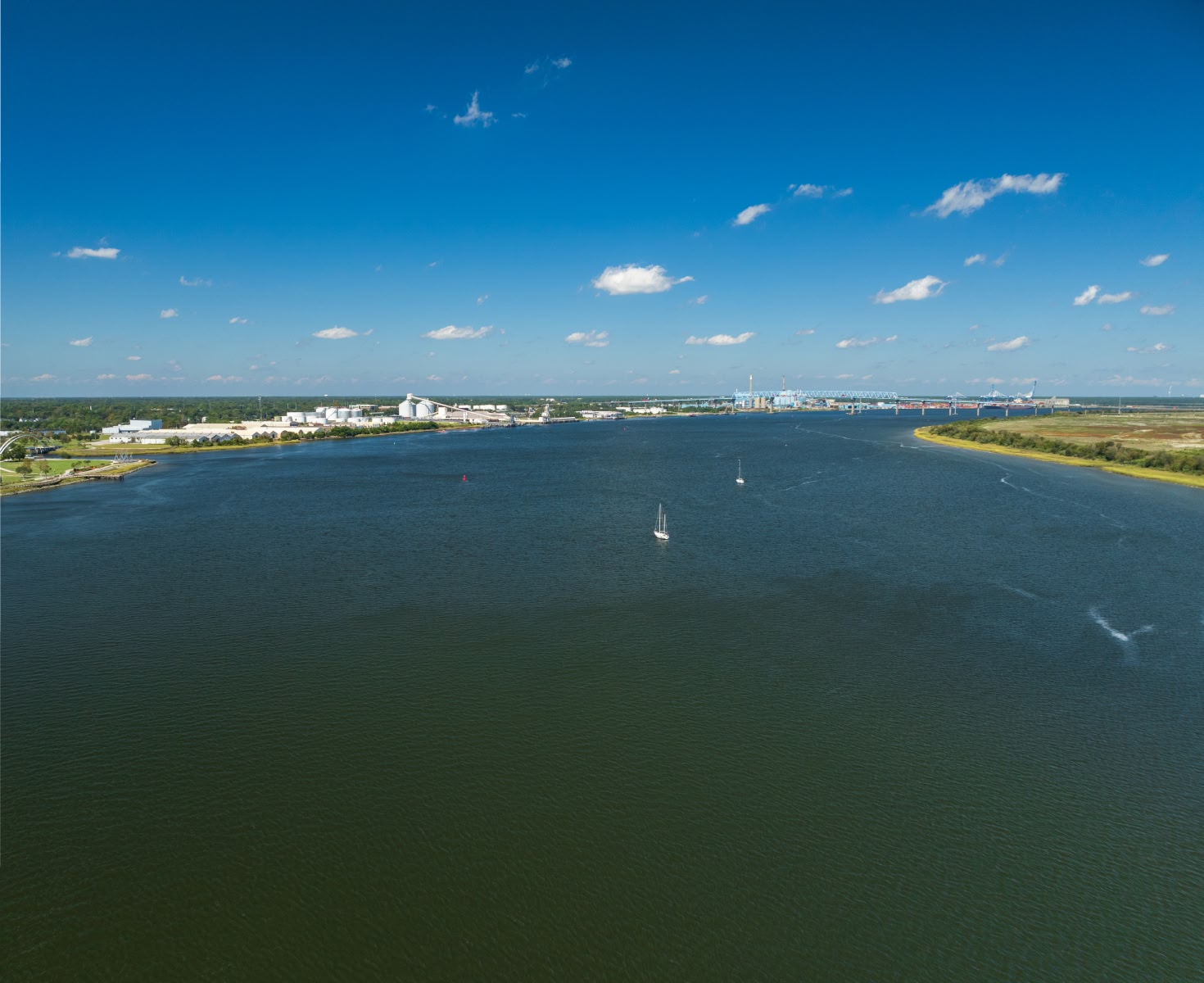 A wide river with deep blue water and two sailboats, bordered by green land. The background features a distant cityscape under a clear sky.