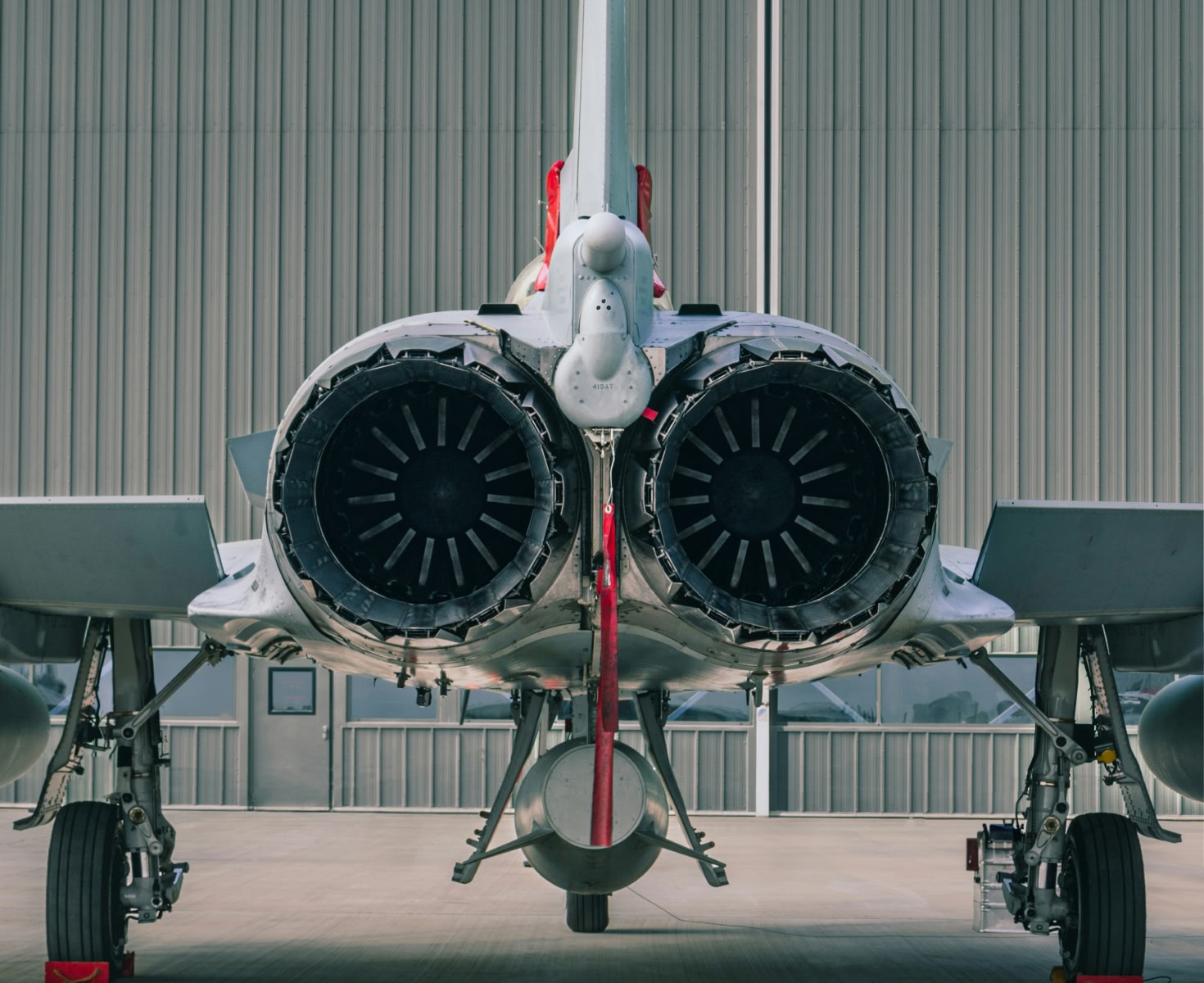 Rear view of a fighter jet on a runway, emphasizing dual exhaust nozzles and landing gear. The backdrop is a large, corrugated hangar door.