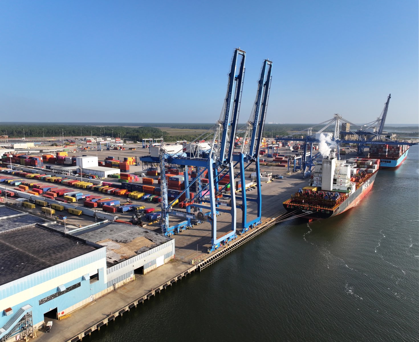 Aerial view of a busy port with docked cargo ships and numerous colorful shipping containers. Tall blue cranes tower over the bustling scene under a clear blue sky.