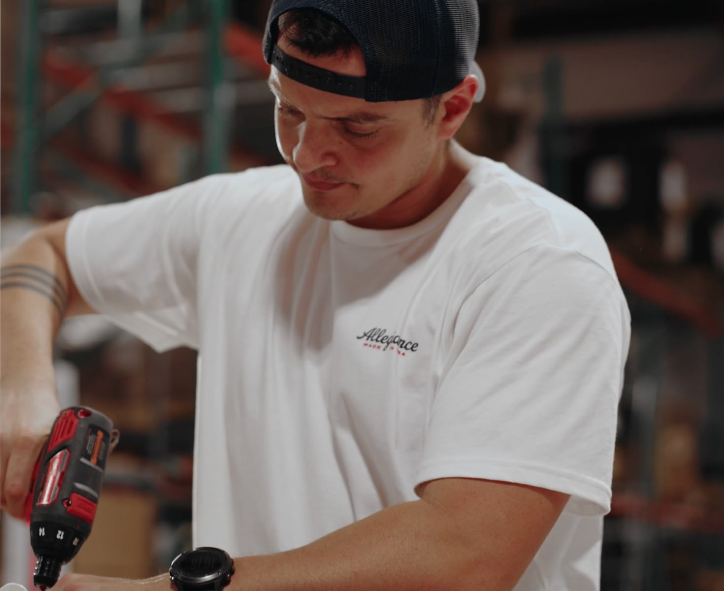 A person in a white t-shirt and black cap uses a red power drill in a workshop. They appear focused, with a blurred industrial background.