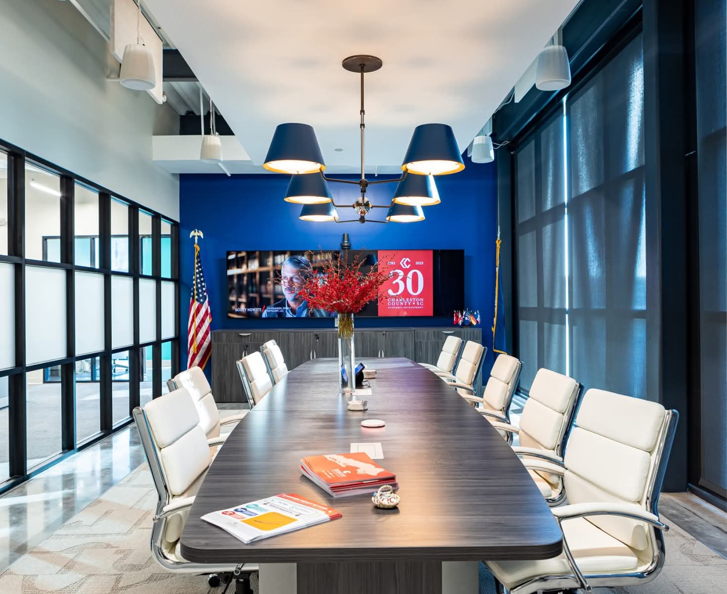 Sleek conference room with a long table, white chairs, and modern pendant lights. A screen displays a 30th anniversary graphic. U.S. flag