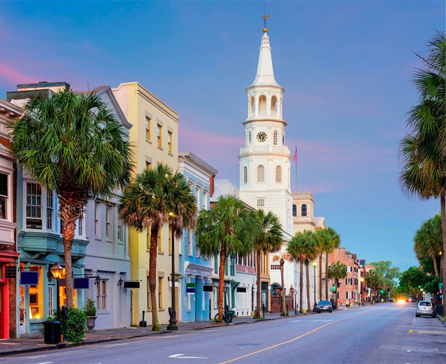 Colorful historic street in Charleston with pastel buildings and palm trees. A white church steeple rises under a vibrant pink and blue sky at dusk.