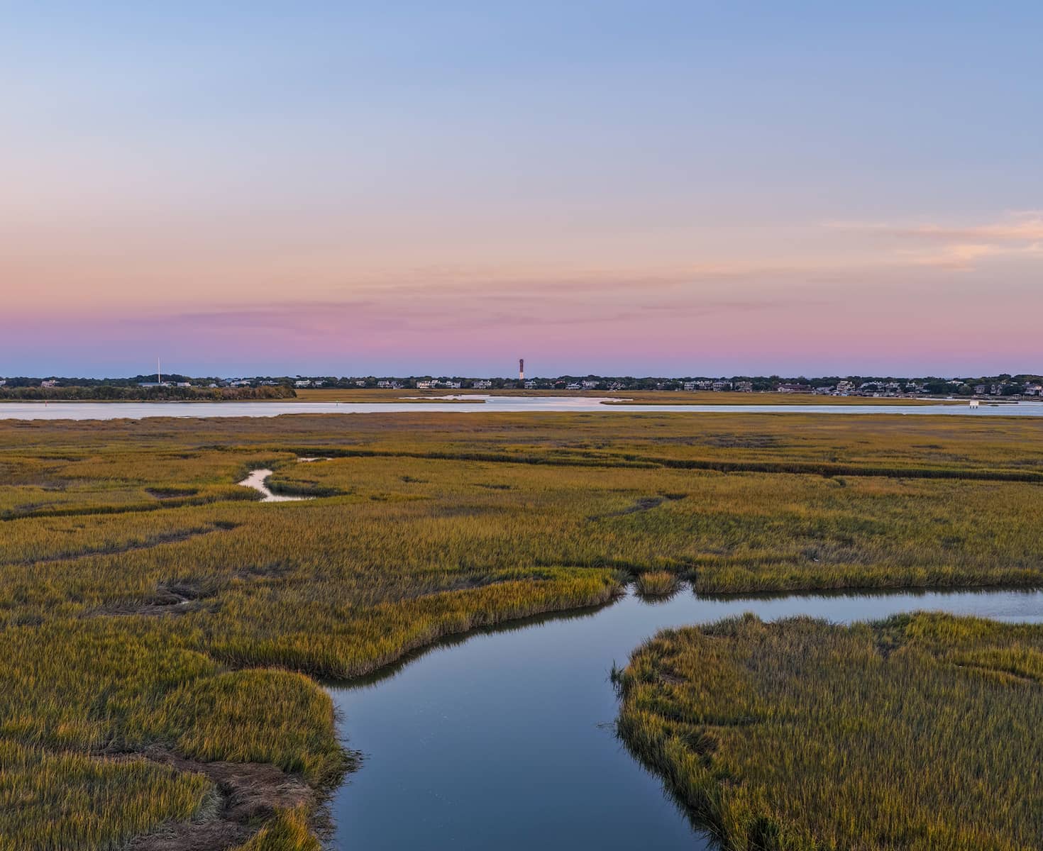 Sunset view of a vast marshland with winding water channels under a pastel sky. In the distance, buildings line the horizon.