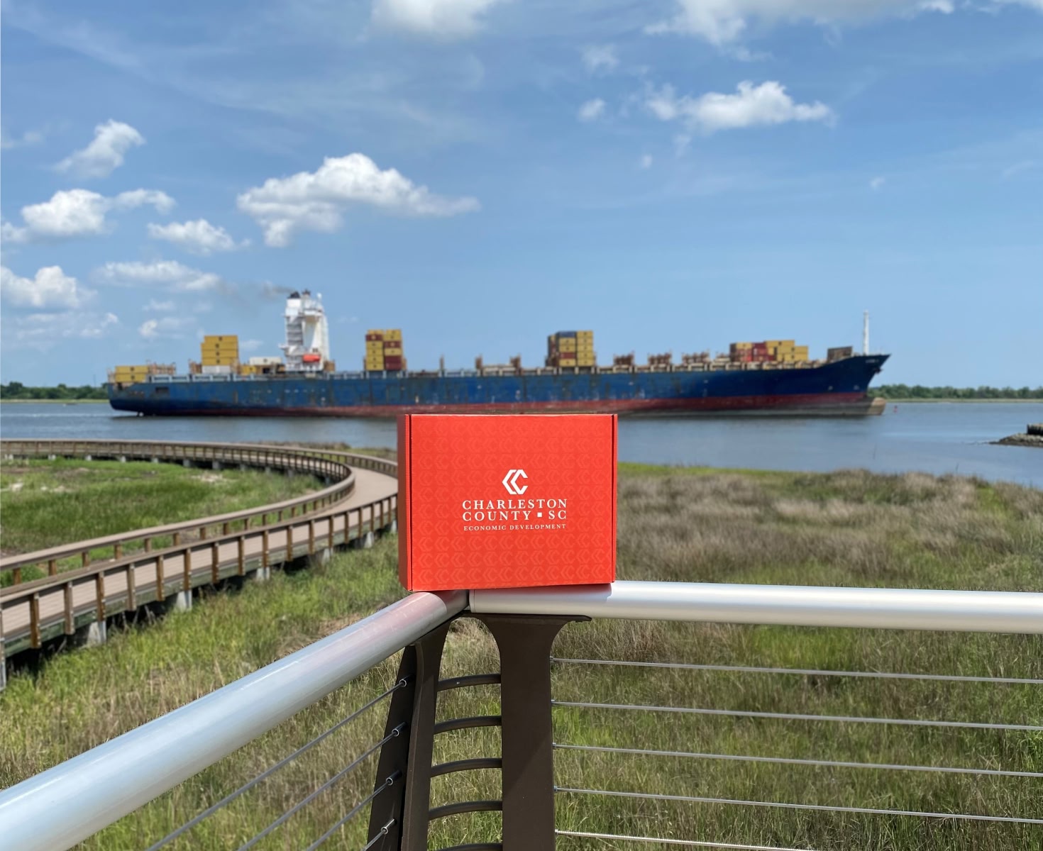 A large cargo ship sails in the background on a calm river under a blue sky. In the foreground, a red Charleston County SC box sits on a railing.
