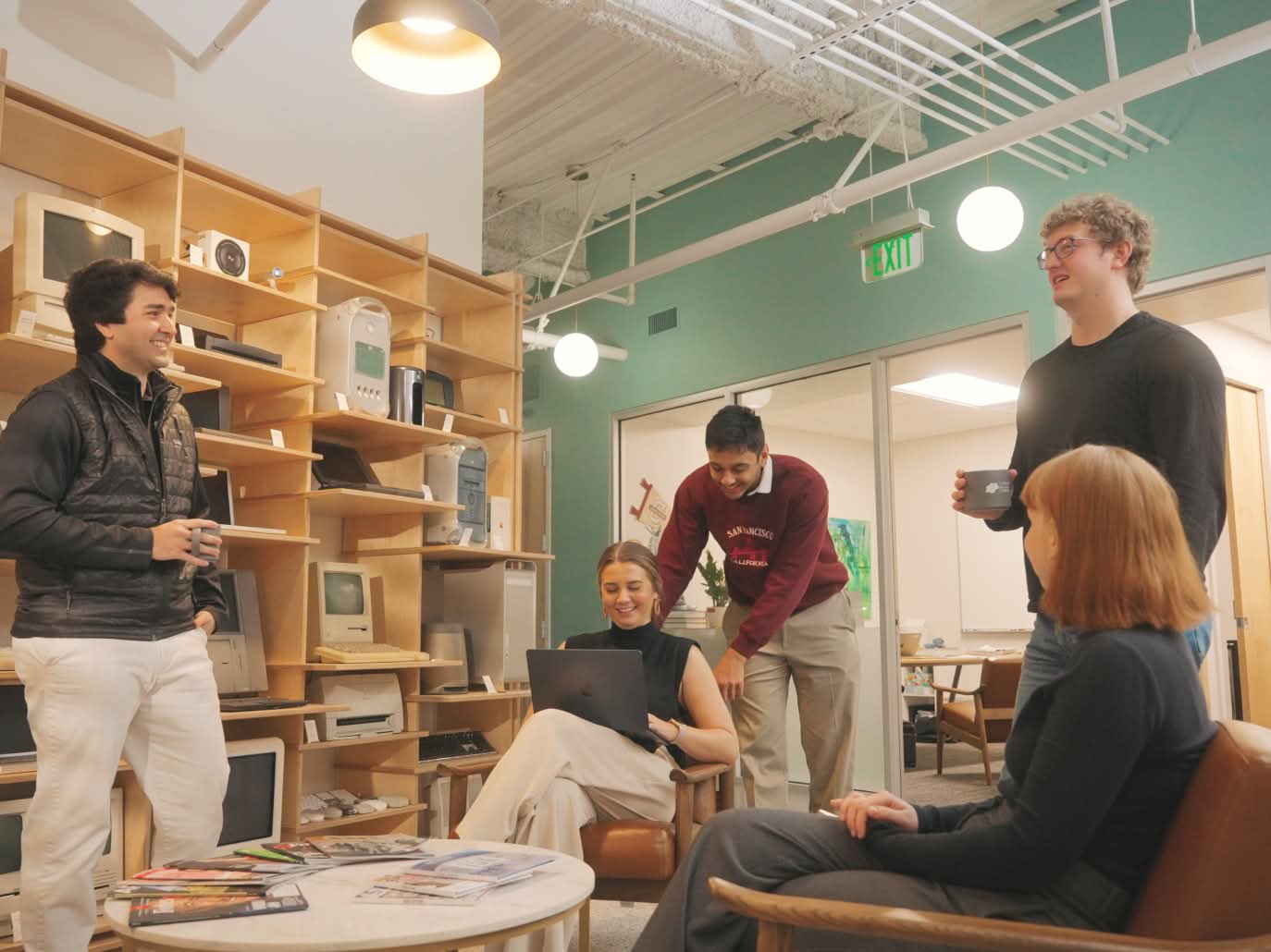 A group of five colleagues in a casual office space shares a laugh. Shelves with vintage electronics line the walls, creating a modern-retro vibe.