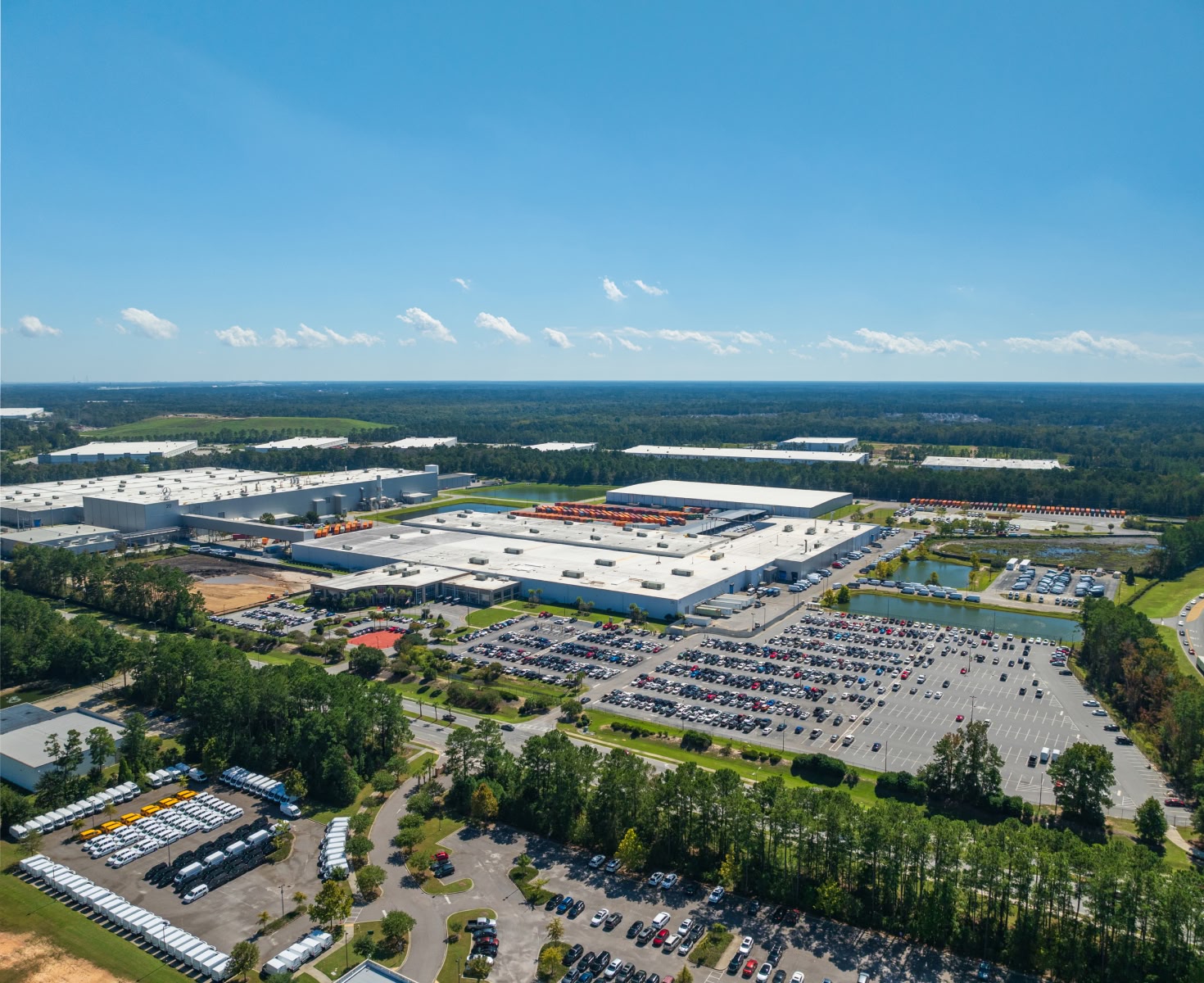 Aerial view of a sprawling industrial complex surrounded by parking lots filled with cars, lush greenery, and trees under a clear blue sky.