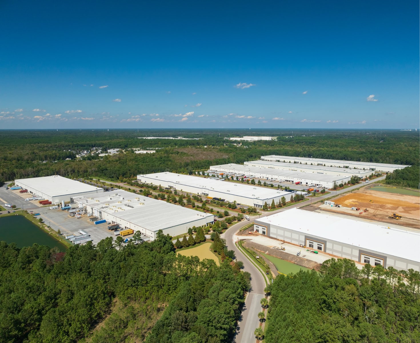 Aerial view showing multiple large white-roofed industrial buildings surrounded by green trees under a clear blue sky