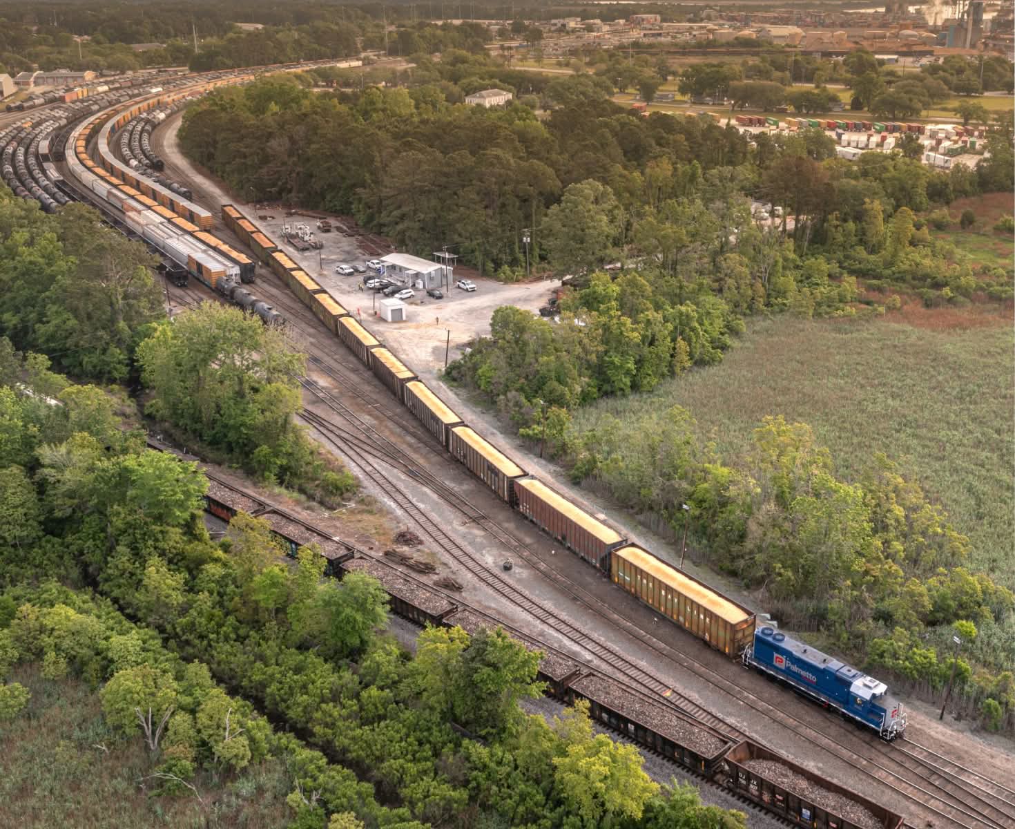 Aerial view of a blue locomotive pulling freight cars on a curved track through a lush, green landscape. Nearby are trees, a small building, and parked trucks, with a hazy background showing a distant town.