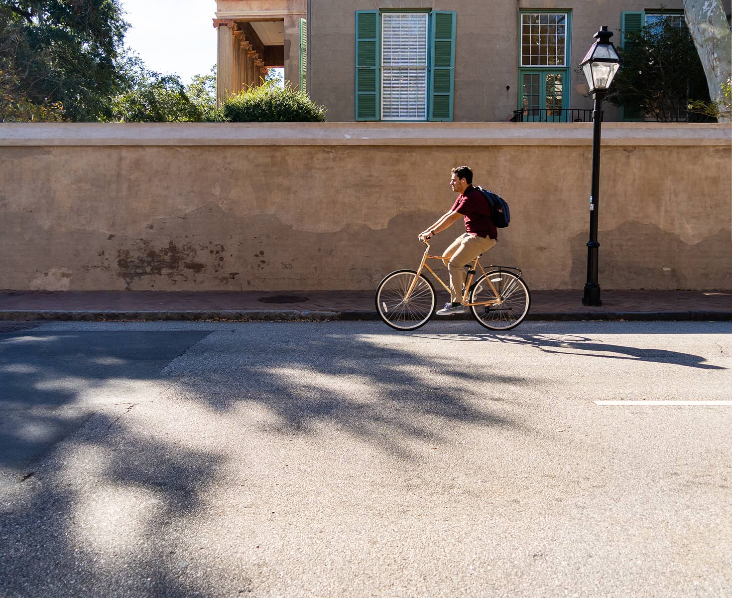 A person rides a bicycle along a sunlit street, next to a beige stucco wall. They wear a red shirt and backpack