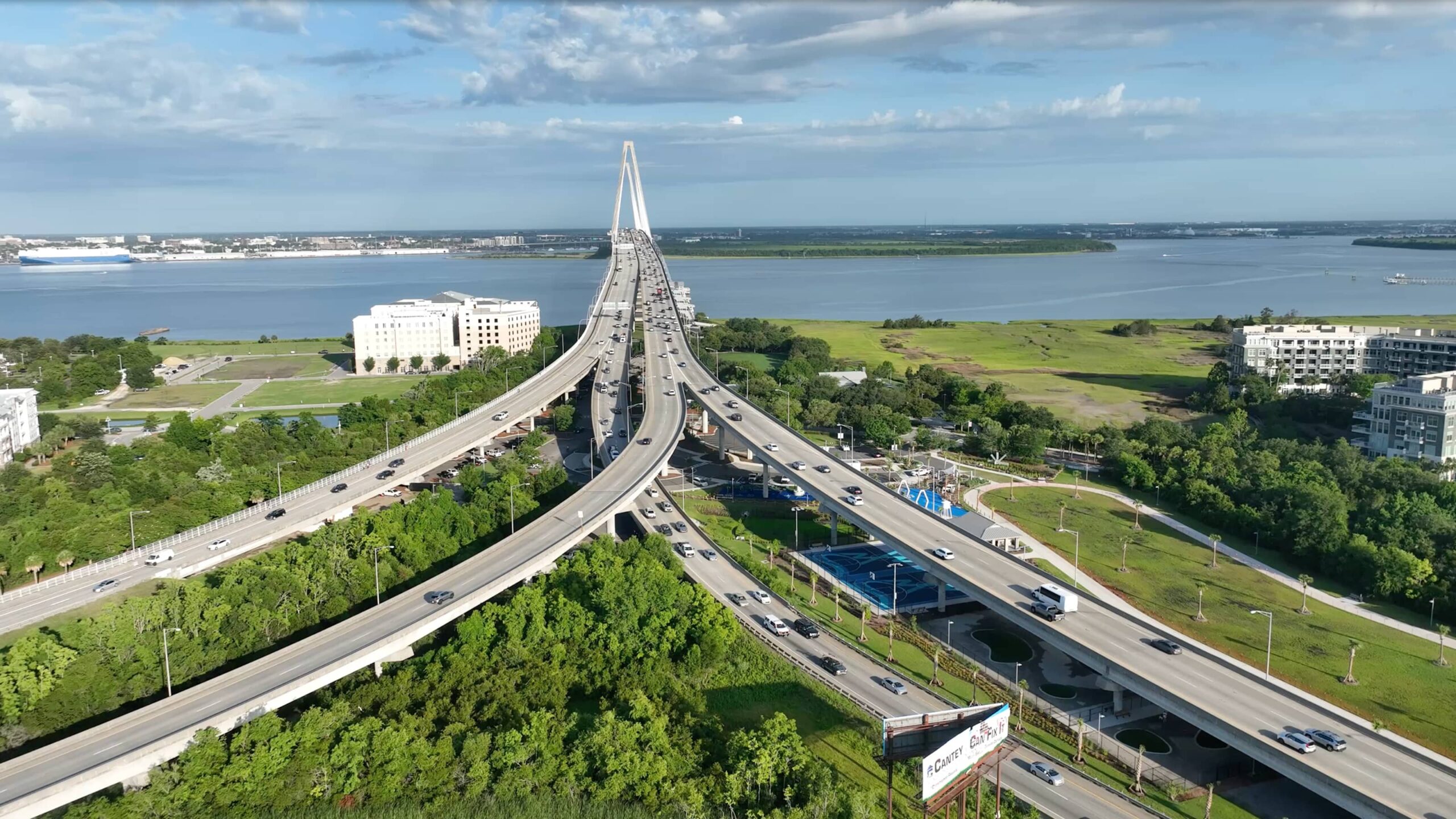 Aerial view of a large cable-stayed bridge spanning a wide river, connecting lush green land. Cars traverse the bridge under a partly cloudy blue sky.