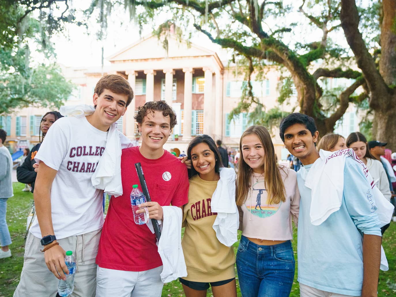 Five smiling young adults pose together outdoors with arms around each other, wearing casual college-themed attire. A historic building and trees are in the background.
