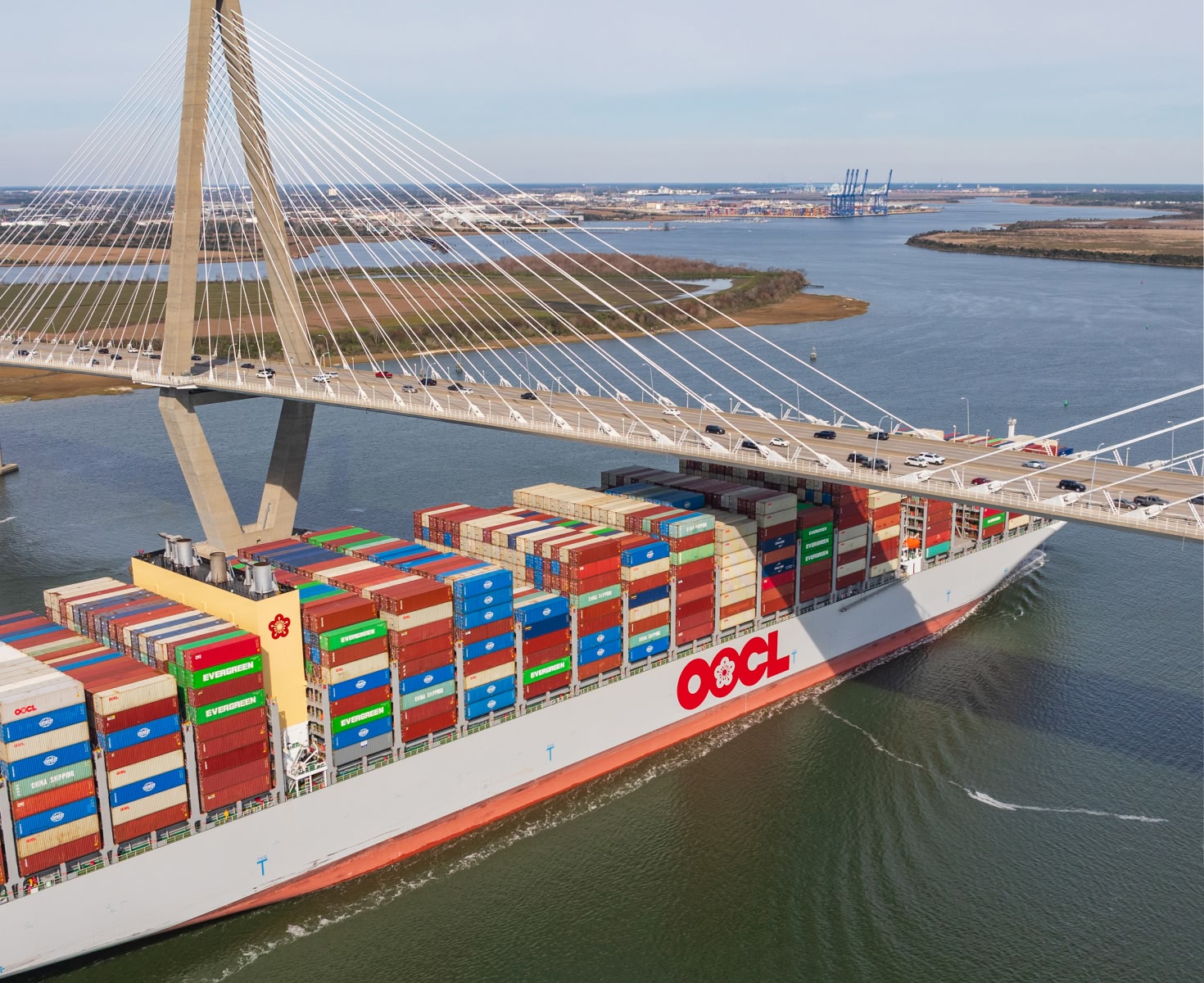 A massive cargo ship with colorful containers passes under a large, modern cable-stayed bridge on a wide river, indicating bustling trade activity.