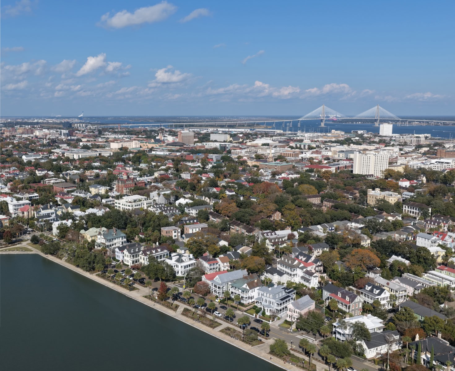 Aerial view of a charming coastal cityscape with historic houses, tree-lined streets, a wide river, and a large bridge under a bright blue sky.