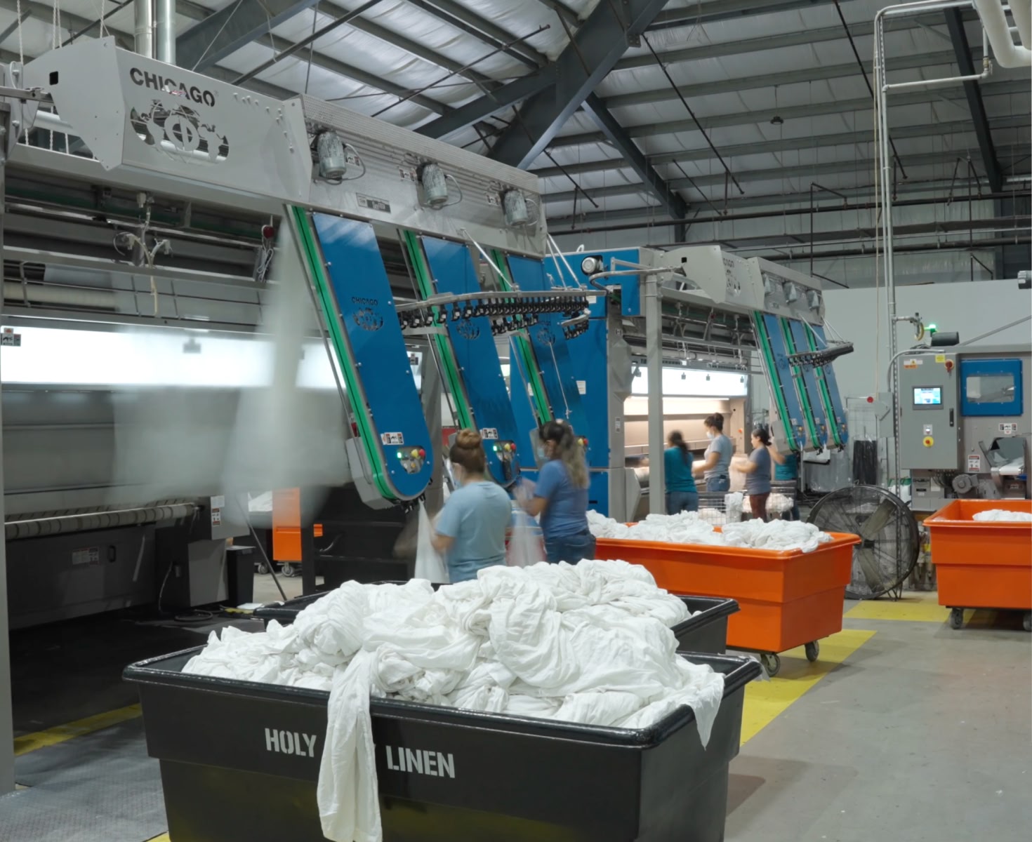 Industrial laundry facility with large machines and bright blue panels. Workers handle white linens, neatly stacked.