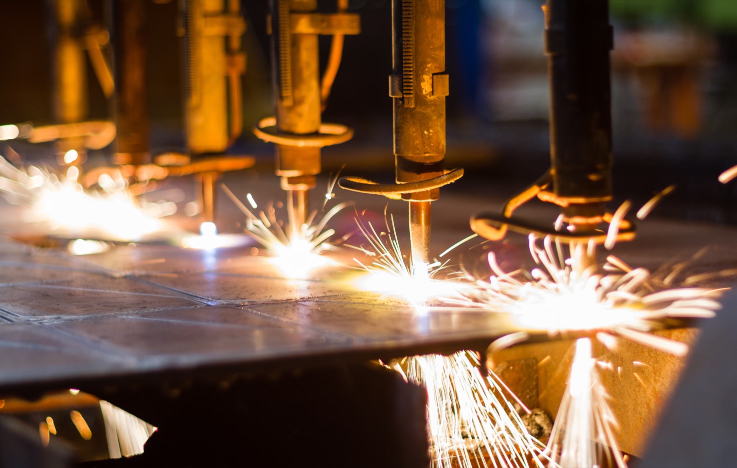 A close-up, perspective view of multiple industrial laser or plasma cutting torches in a row. Bright white and orange sparks fly as the machines cut through a thick metal sheet in a manufacturing facility