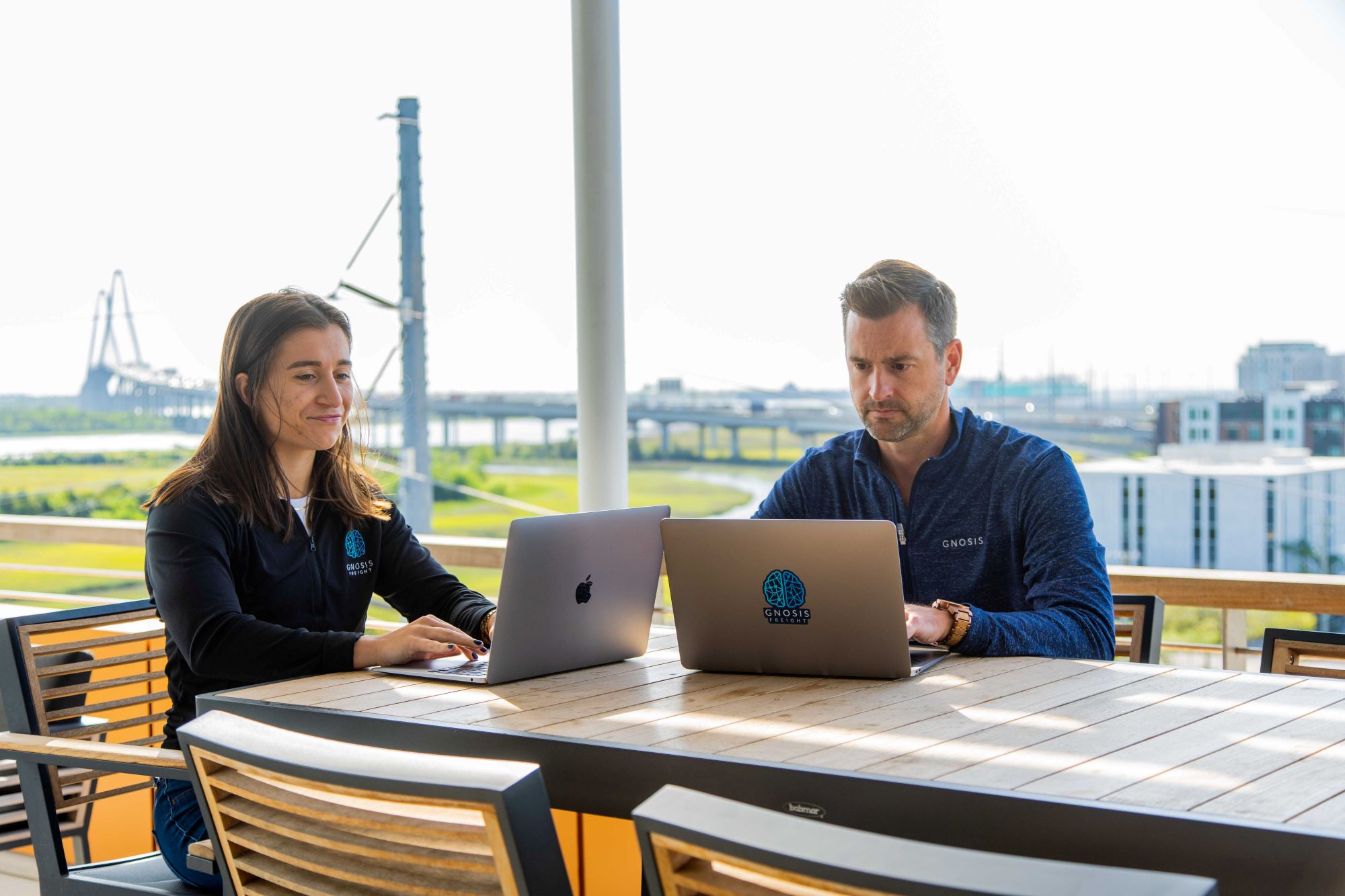 A man and a woman wearing Gnosis Freight branded apparel working on laptops at an outdoor table. They are on a balcony overlooking a scenic view of a large suspension bridge and coastal landscape in Charleston.