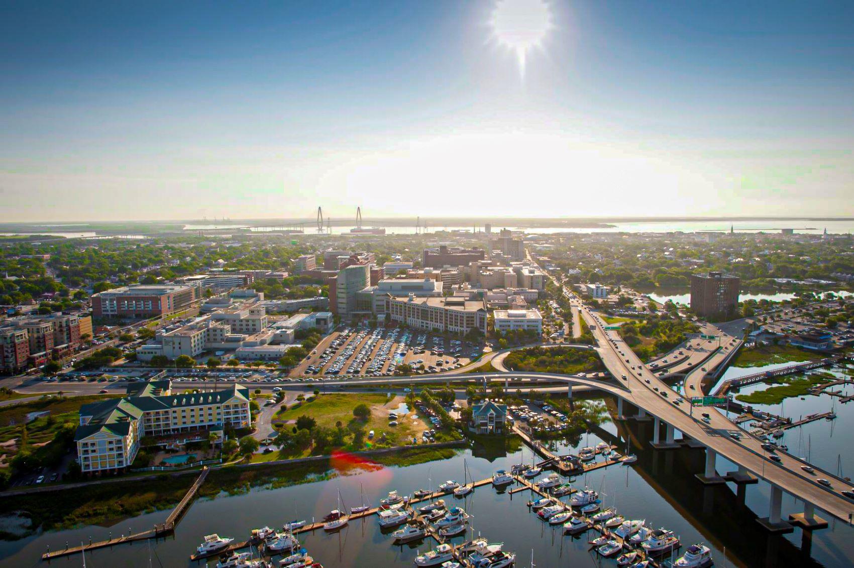 An aerial, high-angle view of the City of Charleston featuring a marina filled with white boats in the foreground and a large highway overpass. The sun is high in a clear sky, illuminating the urban landscape, medical district, various other buildings, and a distant bridge over the water.